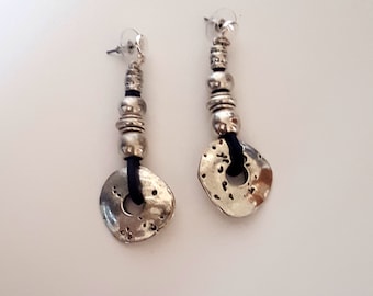 May include: A pair of silver-toned dangle earrings. Each earring features a series of beads and a textured, circular pendant. A black cord connects the pendant to the beaded portion. The earrings have a post-back closure.