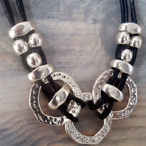 May include: A black leather necklace with silver beads and three large silver rings. The rings are connected and have a textured surface.
