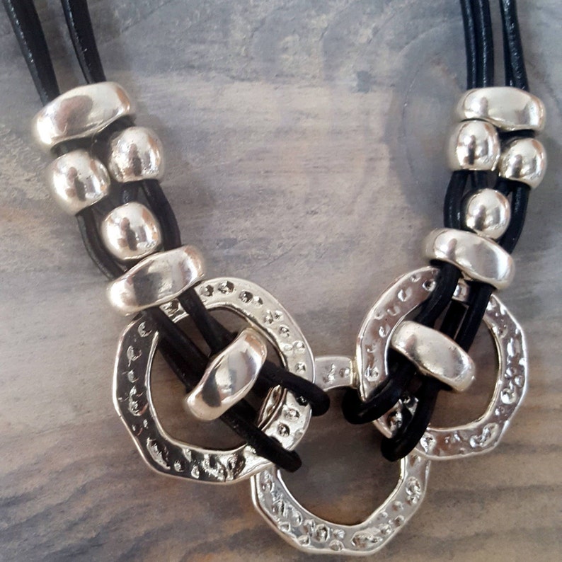 May include: A black leather necklace with silver beads and three large silver rings. The rings are connected and have a textured surface.