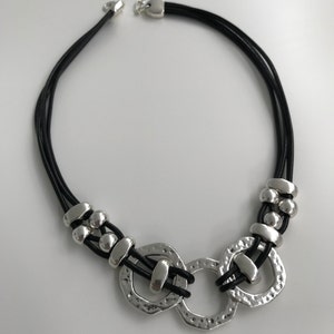 May include: A black leather necklace with three silver rings and silver beads. The rings are textured and have a hammered finish.
