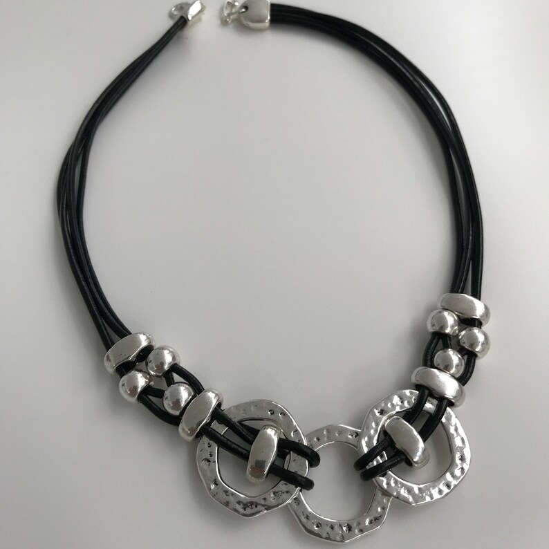 May include: A black leather necklace with three silver rings and silver beads. The rings are textured and have a hammered finish.