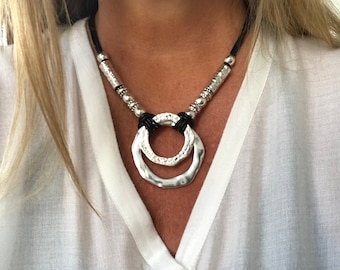 May include: A silver necklace with a large, double-circle pendant and black leather cord. The necklace features silver beads along the cord.