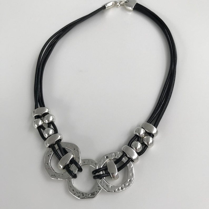 May include: A black leather necklace with three silver rings and silver beads. The rings are connected and have a hammered texture.