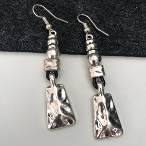May include: Silver dangle earrings with a textured, rectangular pendant and a series of silver beads. The earrings have black leather cord accents.