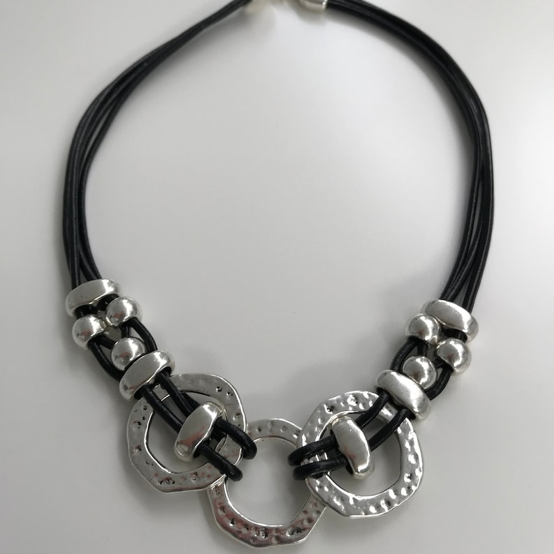 May include: A black leather necklace with three silver rings and silver beads. The rings are textured and have a hammered finish.