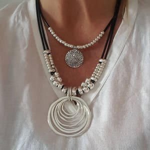 Könnte beinhalten: Eine mehrreihige silberne Halskette mit einem großen, runden Anhänger, der aus mehreren konzentrischen Kreisen besteht. Die Kette besteht aus schwarzem Lederband und silbernen Perlen.