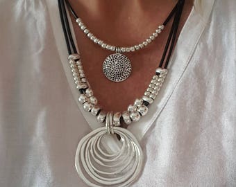 May include: A layered silver necklace with a large, circular pendant featuring multiple concentric circles. The necklace is made of black leather cord and silver beads.