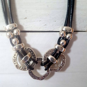 May include: A black leather necklace with silver metal circles and beads. The necklace has a layered design with three strands of leather.