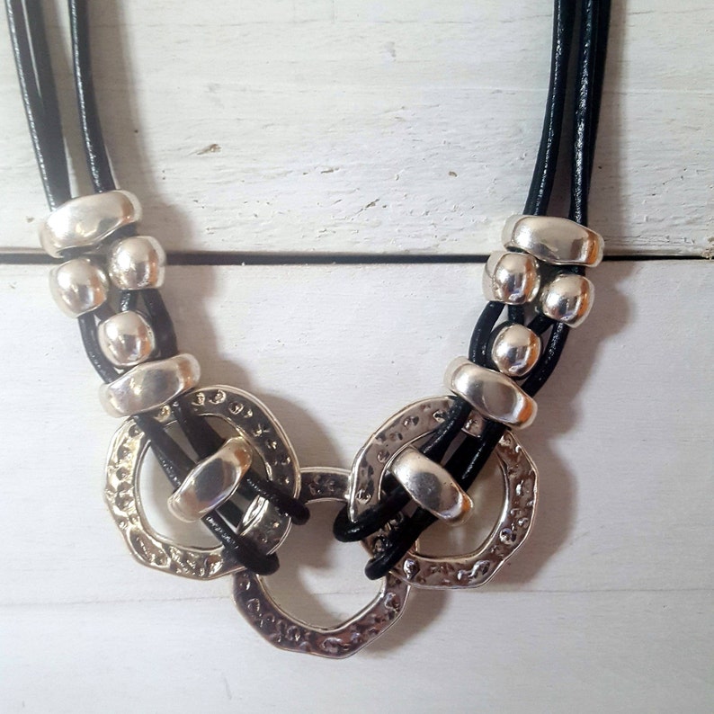 May include: A black leather necklace with silver metal circles and beads. The necklace has a layered design with three strands of leather.