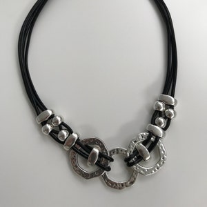 May include: A black leather necklace with three silver rings and silver beads. The rings are textured and have a hammered finish.
