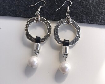 May include: A pair of silver-toned earrings with a hammered texture, featuring a black leather accent and a dangling white pearl. The earrings have a hook closure and a circular design, suitable for everyday wear.