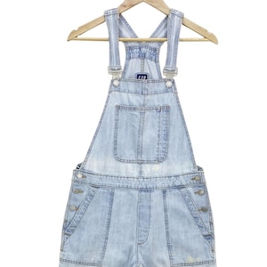 Puede incluir: Pantalón vaquero de mezclilla azul claro con tirantes ajustables y un solo bolsillo delantero. El pantalón tiene un corte holgado y está colgado en una percha de madera.