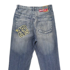 Puede incluir: Un par de pantalones vaqueros azules de mezclilla con un aspecto descolorido. El bolsillo trasero tiene un parche bordado amarillo y negro que dice "AKD MKS".