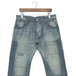 Op de afbeelding: Lichtblauwe denim jeans met een vervaagde wassing en patchdetails. De jeans hangen aan een houten hanger tegen een witte achtergrond. Kenmerken zijn een knoopsluiting, zakken en een rechte pijp.