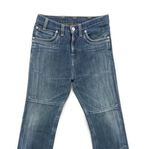 Op de afbeelding: Een paar blauwe denim jeans met een rechte pijp en een knoopsluiting. De jeans heeft een distressed look met verbleekte plekken en stiksels.