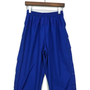 Pode incluir: Calça de treino azul com fecho de botões de pressão nas laterais. A calça tem uma cintura elástica.