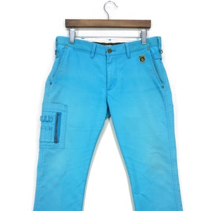 May include: A pair of light blue cargo trousers with a zipped pocket on the left leg. The trousers have a belt loop waistband and a gold patch on the right back pocket.