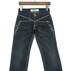 Puede incluir: Vaqueros de mezclilla azul oscuro con detalles desgastados y un botón de metal dorado. Los vaqueros tienen un diseño único con una costura en forma de V en la parte delantera y un pequeño bolsillo en el muslo derecho.