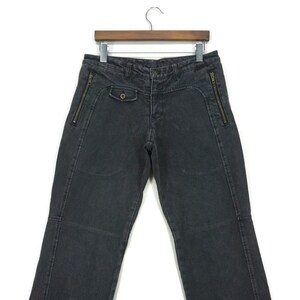 May include: Black denim trousers with zipper details on the sides and a button fastening. The trousers are hanging on a wooden hanger.