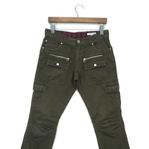 May include: Green cargo pants with multiple pockets and zipper details. The waistband has a red and black plaid lining.