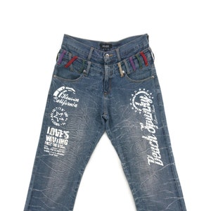Puede incluir: Vaqueros de mezclilla azul con diseños gráficos blancos. Los diseños incluyen "Heaven California", "Love's Waiting" y "Beach Jewelry".