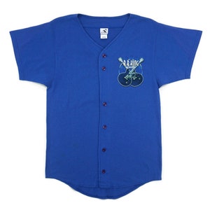 Puede incluir: Una camiseta de béisbol azul con cuello blanco y cierre de botones. La camiseta tiene un gráfico de dos guitarras cruzadas con el texto "B.B. King" en la parte delantera.