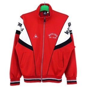 Puede incluir: Chaqueta de chándal roja, blanca y negra con cierre de cremallera. La chaqueta tiene un logotipo de Le Coq Sportif en la manga derecha y el texto "WAYO UNIV SKI TEAM" en la parte delantera.