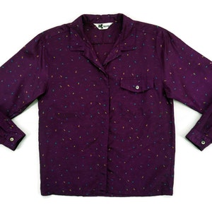 May include: A purple long-sleeved button-down shirt with a small, colorful print. The shirt has a pocket on the left side and a button-up collar.