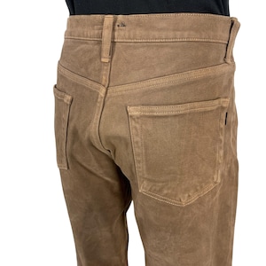 Puede incluir: Primer plano de unos pantalones vaqueros marrones. Los pantalones tienen un diseño clásico de cinco bolsillos con cierre de botón y cremallera. La tela parece ser un denim resistente y duradero, y los pantalones tienen un aspecto ligeramente desgastado.