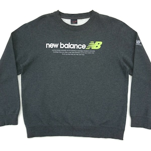 以下が含まれることがあります： 左胸に白の「new balance」ロゴと緑色のニューバランスロゴが付いたダークグレーのスウェットシャツ。長袖でクルーネックです。