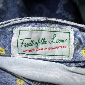Puede incluir: Un primer plano de una etiqueta de mezclilla azul con costuras blancas. La etiqueta dice "Fruit of the Loom" en una fuente de letra cursiva verde y blanca. Debajo del nombre, dice "Unconditionally Guaranteed" en una fuente negra m&aacute;s peque&ntilde;a.
