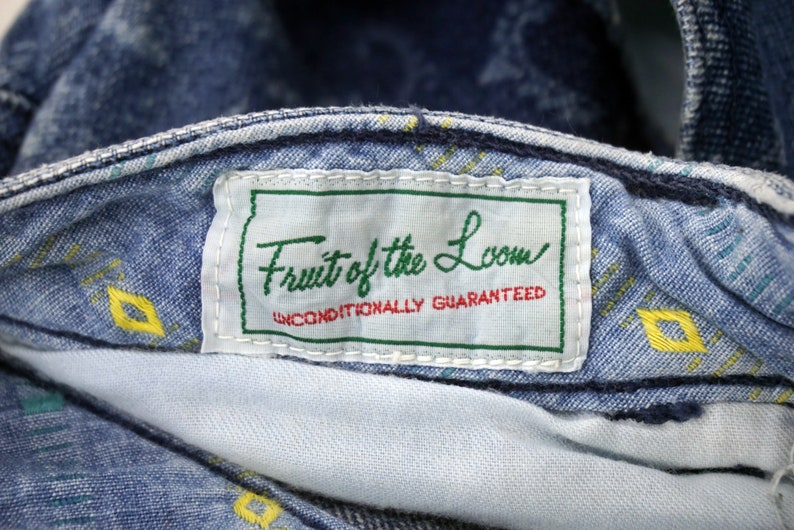 Puede incluir: Un primer plano de una etiqueta de mezclilla azul con costuras blancas. La etiqueta dice "Fruit of the Loom" en una fuente de letra cursiva verde y blanca. Debajo del nombre, dice "Unconditionally Guaranteed" en una fuente negra m&aacute;s peque&ntilde;a.