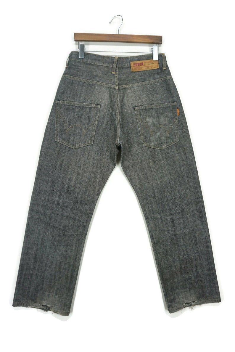 May include: A pair of dark grey denim jeans with a distressed hem. The jeans have a faded look and a small orange tag on the back pocket. The jeans have the word "EDWIN" on a brown label on the back pocket.