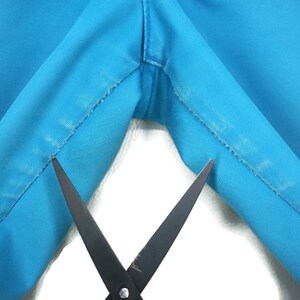 May include: Close-up of a pair of scissors cutting a seam on a bright blue fabric. The fabric is folded in half, and the scissors are positioned in the middle of the fold.