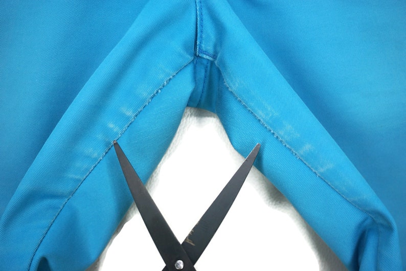 May include: Close-up of a pair of scissors cutting a seam on a bright blue fabric. The fabric is folded in half, and the scissors are positioned in the middle of the fold.