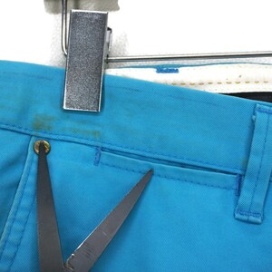 May include: A pair of blue denim trousers with a single pocket on the right side. The trousers have a dark brown leather belt loop and a small metal button.