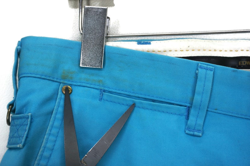 May include: A pair of blue denim trousers with a single pocket on the right side. The trousers have a dark brown leather belt loop and a small metal button.