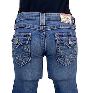 Könnte beinhalten: Nahaufnahme einer blauen Jeans mit dekorativen Nähten und Gesäßtaschen. Die Jeans hat einen True Religion Markenaufnäher und Knöpfe. Die Nähte sind in einer kontrastierenden gelben Farbe.