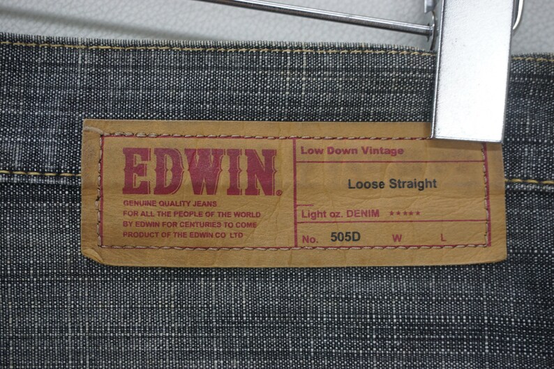 May include: A brown leather label with the text "EDWIN" in red letters. The label also includes the text "Low Down Vintage", "Loose Straight", "Light oz. DENIM", "No. 505D", "W", and "L".