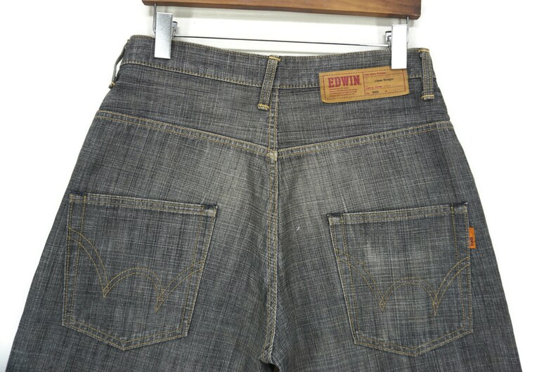 May include: A pair of dark grey denim jeans with a brown leather label that reads "EDWIN" on the back waistband. The jeans have two back pockets with yellow stitching detail.