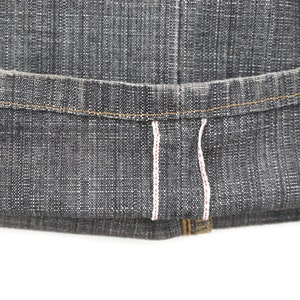 May include: Close-up of the hem of a pair of dark grey denim jeans. The jeans have a raw hem with white stitching and a red accent.