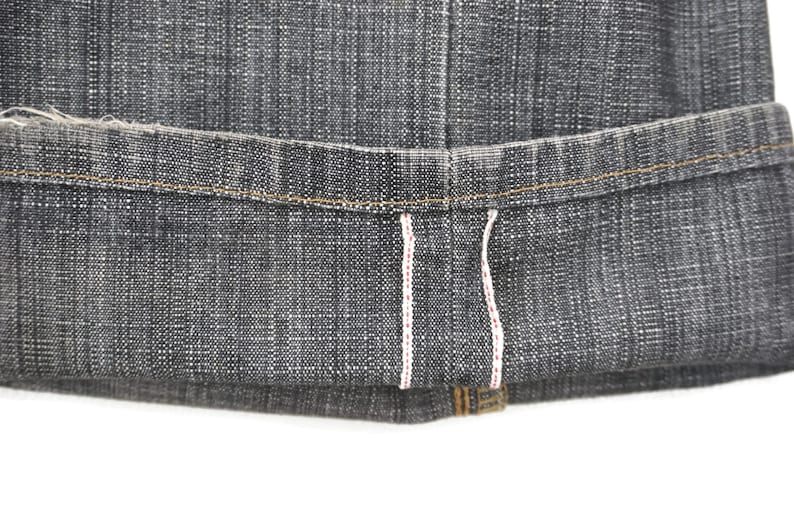 May include: Close-up of the hem of a pair of dark grey denim jeans. The jeans have a raw hem with white stitching and a red accent.