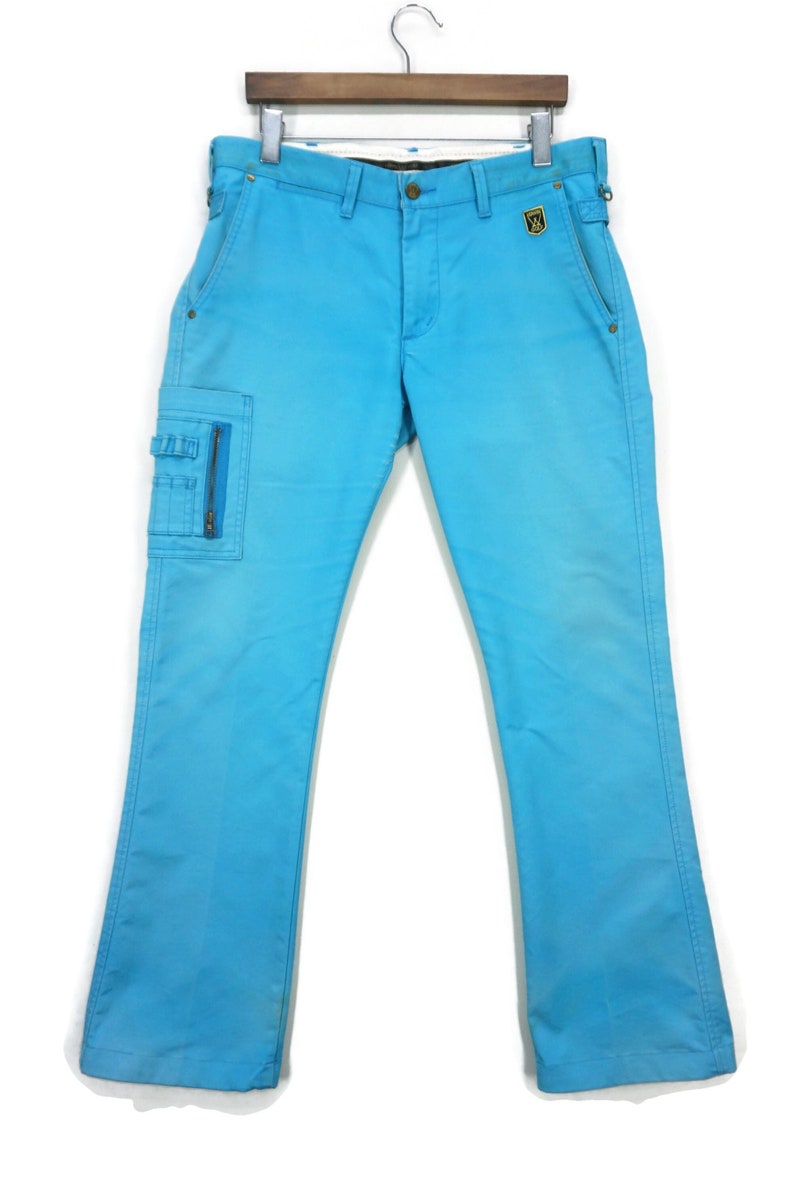 May include: A pair of light blue cargo trousers with a zipped pocket on the left leg. The trousers have a belt loop waistband and a gold patch on the right back pocket.