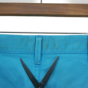 May include: A pair of blue trousers with a black pair of scissors on top. The trousers have a belt loop and a pocket on the right side.