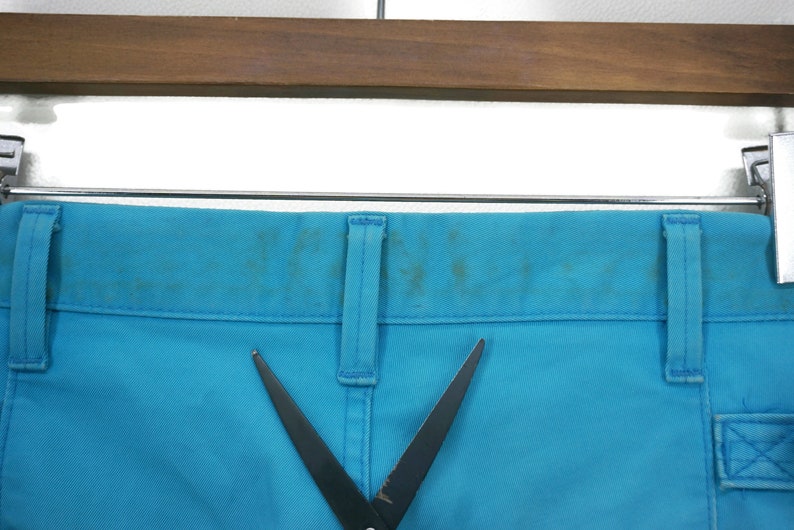 May include: A pair of blue trousers with a black pair of scissors on top. The trousers have a belt loop and a pocket on the right side.