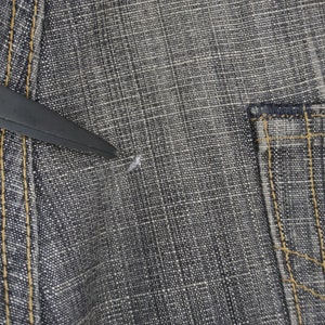 May include: Close-up of a pair of dark grey denim jeans with a small hole in the fabric. The jeans have a pocket on the right side and a seam down the left side.