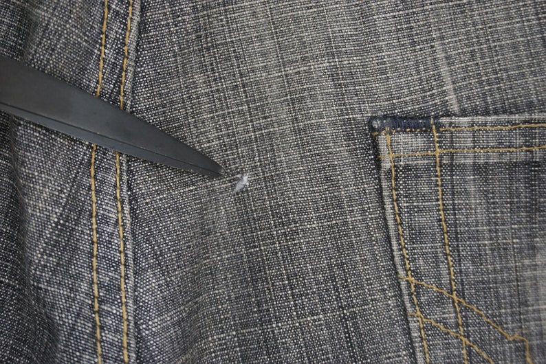 May include: Close-up of a pair of dark grey denim jeans with a small hole in the fabric. The jeans have a pocket on the right side and a seam down the left side.