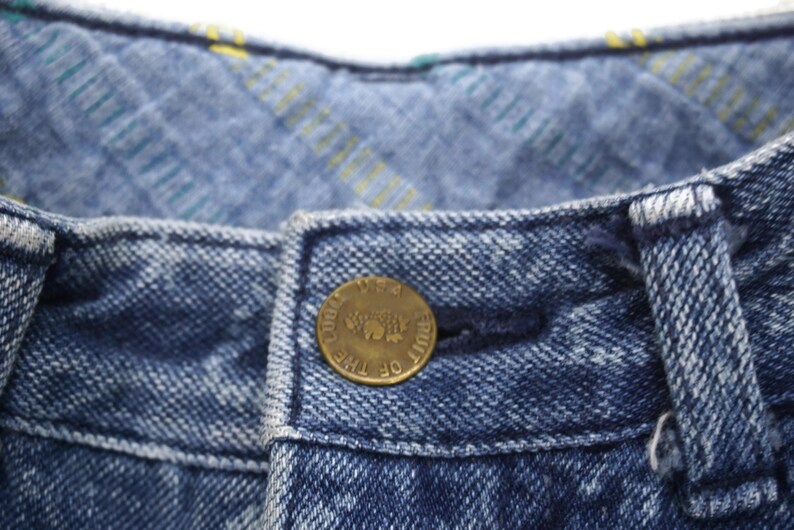 Puede incluir: Primer plano de una cintura de mezclilla azul con un bot&oacute;n de lat&oacute;n que dice "Fruit of the Loom" en un dise&ntilde;o circular.