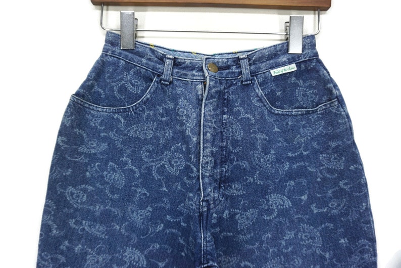 Puede incluir: Vaqueros azules de mezclilla con estampado paisley. Los vaqueros tienen cintura alta y cierre de bot&oacute;n. La etiqueta de los vaqueros dice "Paris of the Lane".
