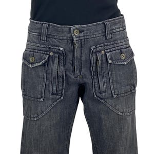 Puede incluir: Pantalones vaqueros grises oscuros con múltiples bolsillos delanteros y cierre de botón y cremallera. Los pantalones tienen un diseño único con varios bolsillos y una apariencia descolorida, lo que sugiere un aspecto desgastado.